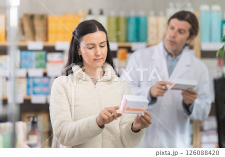 Middle-aged woman choosing pills in box while pharmacist offering in drugstore Middle-aged woman choosing pills in box while pharmacist offering in drugstore 126088420
