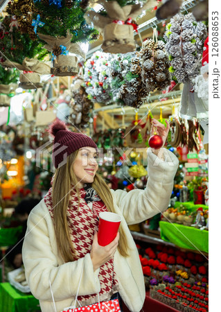 Young girl customer with glass of coffee in her hands stopped near showcase with Christmas tree toys Young girl customer with glass of coffee in her hands stopped near showcase with Christmas tree toys 126088653