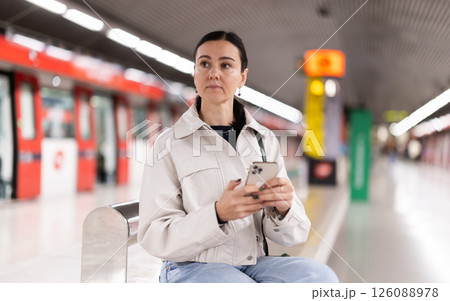 Woman monitors navigation of train in smartphone sitting on a bench at metro station 126088978