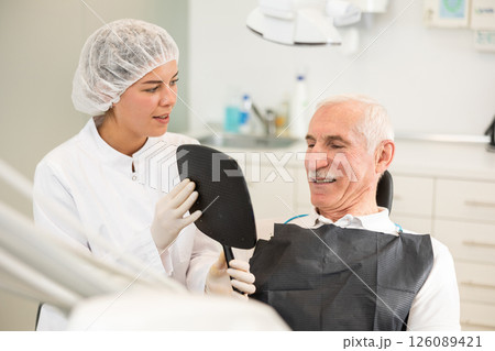 Girl specialist holds mirror in front of senior man patient and suggests evaluating effect of treatment 126089421