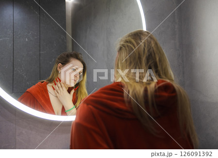 Blonde young woman in a red hoodie with long red nails examines her face in a bathroom mirror with a concerned and focused expression. 126091392
