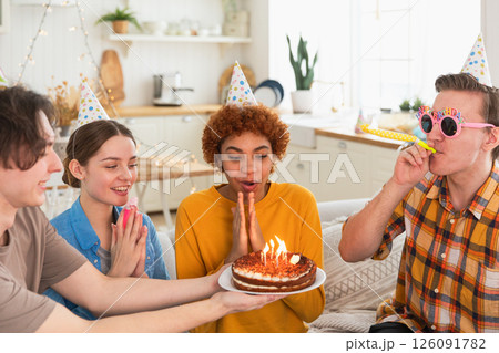Make a wish. Woman wearing party cap blowing out burning candles on birthday cake. Happy Birthday party. Group of friends wishes girl happy birthday. People celebrating birthday with party at home Make a wish. Woman wearing party cap blowing out burning candles on birthday cake. Happy Birthday party. Group of friends wishes girl happy birthday. People celebrating birthday with party at home 126091782