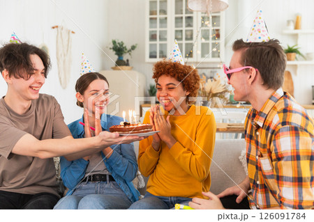 Make a wish. Woman wearing party cap blowing out burning candles on birthday cake. Happy Birthday party. Group of friends wishes girl happy birthday. People celebrating birthday with party at home 126091784