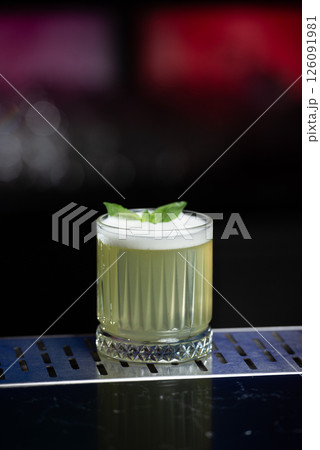 A pale yellow cocktail with a frothy white top is garnished with two vibrant green mint leaves, sitting on a reflective bar surface. 126091981