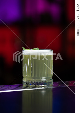 A pale yellow cocktail with a frothy white top is garnished with two vibrant green mint leaves, sitting on a reflective bar surface. 126091982