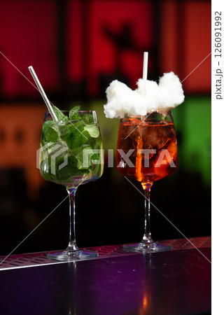 Two uniquely garnished cocktails on a reflective bar surface, one brimming with fresh mint and a straw, the other a vibrant orange hue topped with fluffy white sugar cotton candy and a white straw 126091992