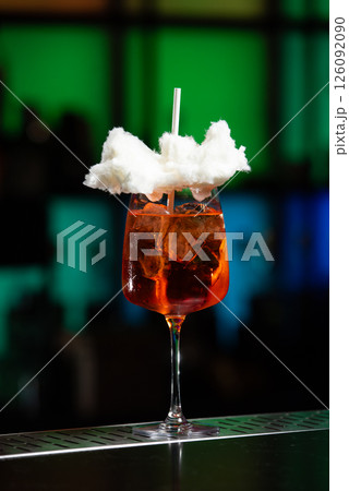 A vibrant orange cocktail filled with ice sits on a textured bar, topped with a whimsical cloud of white cotton candy and a white straw. 126092090