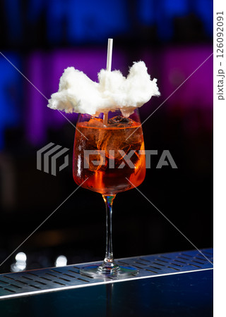 A vibrant orange cocktail filled with ice sits on a textured bar, topped with a whimsical cloud of white cotton candy and a white straw. 126092091