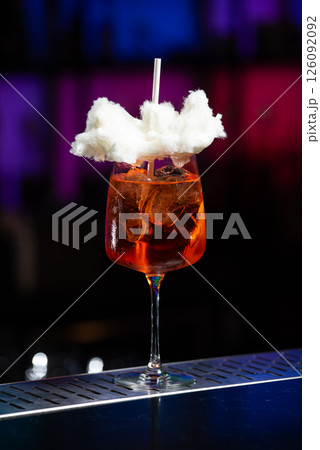 A vibrant orange cocktail filled with ice sits on a textured bar, topped with a whimsical cloud of white cotton candy and a white straw. 126092092