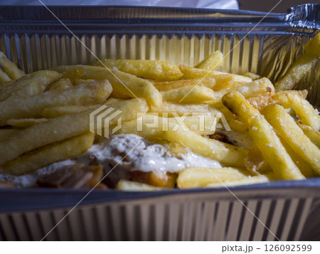 Street food delicacy featuring crispy fries layered over juicy kebab meat, smothered in creamy garlic sauce, served in disposable aluminum container for convenient dining 126092599