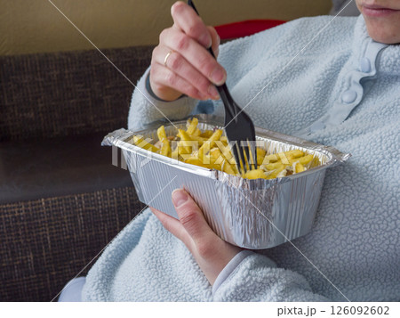 Cozy woman relaxing in pajamas, munching kebab and fries from takeout container while lounging on living room sofa Cozy woman relaxing in pajamas, munching kebab and fries from takeout container while lounging on living room sofa 126092602
