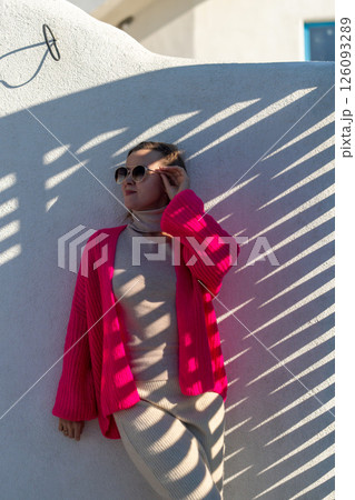 Here's a title attempting to follow all instructions: Cardigan Fashion Greece Summer: Woman poses in pink cardigan against whitewashed wall, showcasing summer style in Greece. 126093289
