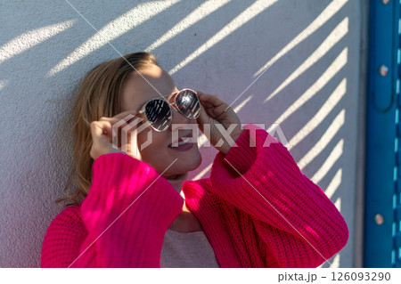 Woman Sunglasses Pink Cardigan: Outdoor sunny portrait, stylish woman adjusts heart-shaped sunglasses, daytime fashion shoot, bright clothing. 126093290