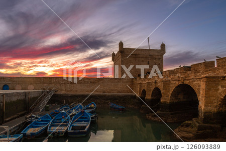 Blue fishing boats in a harbor with foretress bridge and Port of Essaouira Sqala, Portuguese castle in a sunset lights, Essaouira, Morocco 126093889