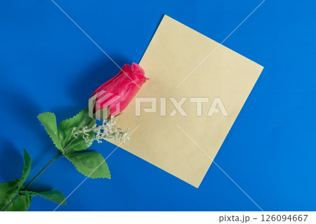 Red rose and blank yellow paper on a blue background for messages or greetings. 126094667