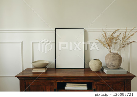 Elegant living room. Mahogany wooden cabinet dresser. Empty picture mock up poster frame. Clay vases, dry grass bouquet, ceramic bowl. Modern Scandinavian interior design, white wall background. Elegant living room. Mahogany wooden cabinet dresser. Empty picture mock up poster frame. Clay vases, dry grass bouquet, ceramic bowl. Modern Scandinavian interior design, white wall background. 126095721