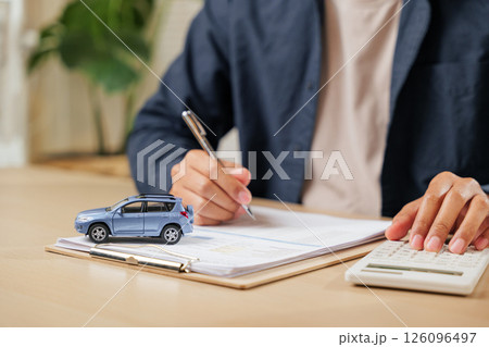 Hands writing a signature on car lease documents while calculating taxes. Financial agreements for loans, contracts, and transactions in buying or selling vehicles. Hands writing a signature on car lease documents while calculating taxes. Financial agreements for loans, contracts, and transactions in buying or selling vehicles. 126096497