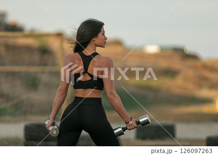 dedicated young woman exercises outdoors while holding dumbbells. She wears a stylish workout outfit and stands confidently showing her strong physique against a scenic background. 126097263