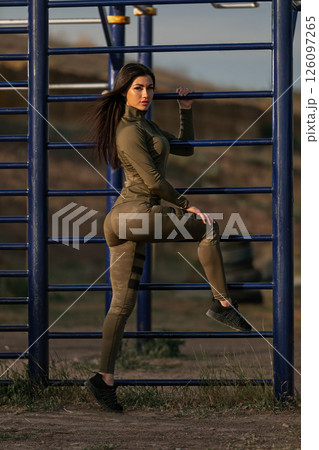 woman wearing a form fitting workout outfit poses confidently by outdoor fitness equipment at sunset showcasing her athleticism and strength in a picturesque setting. 126097265