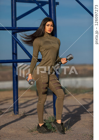 confident woman showcases her fitness style as she stands holding dumbbells at an outdoor gym surrounded by mountains. The clear blue sky adds to the vibrant atmosphere. 126097273