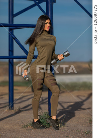 woman in olive green athletic attire holds dumbbells while standing confidently outdoors. The background features blue workout equipment and the day is bright and clear. 126097275