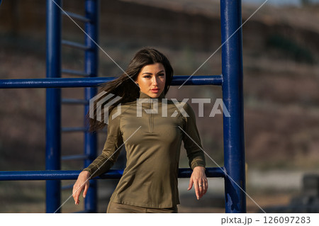 stylish woman in an olive green outfit stands against blue fitness equipment showcasing her confident pose in a sunny outdoor environment. The serene landscape enhances the moment. 126097283