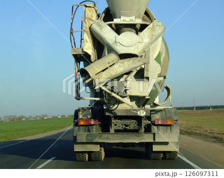 A large concrete mixer truck drives down a rural road, showcasing its heavy machinery and equipment used for construction, with fields visible in the background. A large concrete mixer truck drives down a rural road, showcasing its heavy machinery and equipment used for construction, with fields visible in the background. 126097311