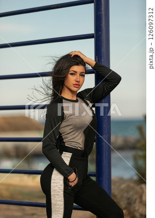 fitness enthusiast demonstrates strength and poise standing next to a blue structure by the ocean. She wears sporty clothing and showcases a vibrant outdoor setting under clear skies. 126097312