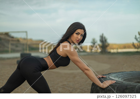 woman performs strength training by pushing a large tire on a fitness field during sunset highlighting her athleticism and outdoor workout routine. woman performs strength training by pushing a large tire on a fitness field during sunset highlighting her athleticism and outdoor workout routine. 126097325
