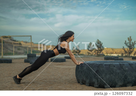 woman in athletic wear exercises by doing push ups on a large tire in an outdoor fitness area during sunset. The sky is partly cloudy and the atmosphere is energetic. woman in athletic wear exercises by doing push ups on a large tire in an outdoor fitness area during sunset. The sky is partly cloudy and the atmosphere is energetic. 126097332