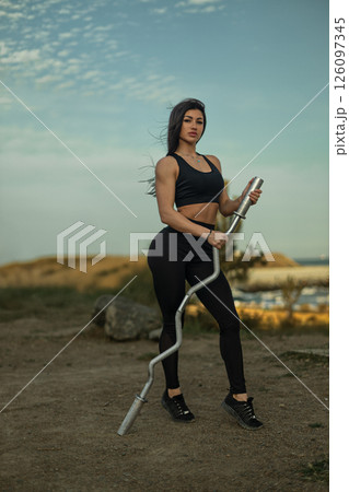 woman showcasing strength stands on a rugged terrain holding a barbell with a scenic coastline in the background. The sunlight adds a warm glow to the scene. 126097345