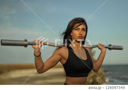 determined woman is lifting a barbell over her shoulders by the shoreline. The sun is setting casting a warm glow on the scene emphasizing her strength and focus. 126097349
