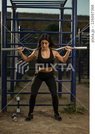woman is engaged in a squat exercise with a barbell at an outdoor gym. The activity takes place in the late afternoon with minimal equipment in sight. woman is engaged in a squat exercise with a barbell at an outdoor gym. The activity takes place in the late afternoon with minimal equipment in sight. 126097366