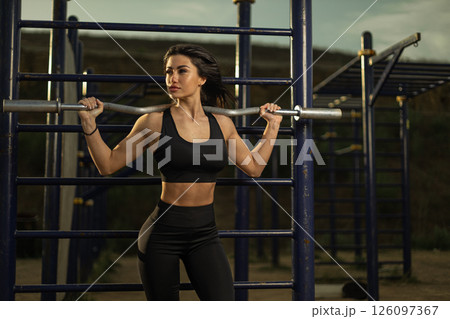 woman in athletic wear confidently lifts a barbell during a strength training session at an outdoor gym. The sun sets in the background creating a warm ambiance. woman in athletic wear confidently lifts a barbell during a strength training session at an outdoor gym. The sun sets in the background creating a warm ambiance. 126097367
