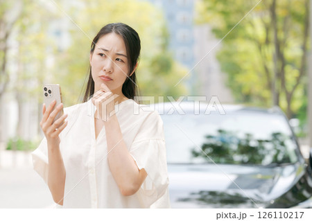 車の前でスマホを見て悩む女性 126110217