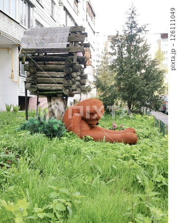 Sad plush bear sitting under a wooden fairy tale house in the yard, surrounded by tall green grass. Unusual scene with emotional undertones 126111049