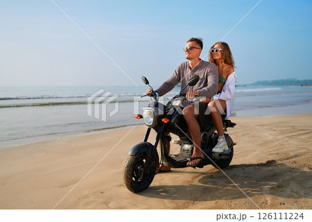 Couple enjoys beach ride on motorbike. Man drives motorcycle with woman on back. Ocean, sand, summer, sunglasses. Adventure travel, romantic journey, tropical holiday by sea. 126111224