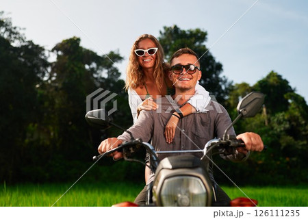 Smiling couple rides motorcycle through tropical jungle. Young man drives motorbike, girlfriend enjoys ride, exploring exotic budget travel. Adventure seekers on scooter. Affordable vacation. Smiling couple rides motorcycle through tropical jungle. Young man drives motorbike, girlfriend enjoys ride, exploring exotic budget travel. Adventure seekers on scooter. Affordable vacation. 126111235