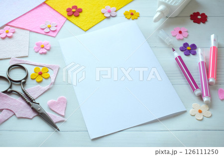 Creative flat lay composition of DIY greeting card crafting materials, including colorful felt flowers, scissors, glue, and blank paper, arranged on a white wooden background, showcasing the process Creative flat lay composition of DIY greeting card crafting materials, including colorful felt flowers, scissors, glue, and blank paper, arranged on a white wooden background, showcasing the process 126111250