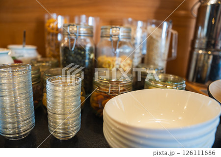 Neatly arranged glass jars with dried fruits, nuts, and cereals on a wooden surface, with glasses, bowls, and a metallic dispenser in the foreground. Neatly arranged glass jars with dried fruits, nuts, and cereals on a wooden surface, with glasses, bowls, and a metallic dispenser in the foreground. 126111686
