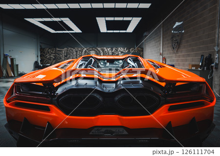 The image captures the rear end of a Lamborghini Revuelto indoors, showcasing its vibrant orange color, angular taillights, and distinctive exhaust system. 126111704