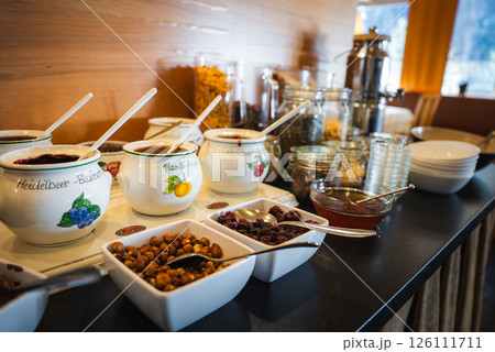 Breakfast buffet features labeled fruit jars, nuts, dried fruits, and cereals. Wooden decor and window reveal an alpine mountain landscape. 126111711
