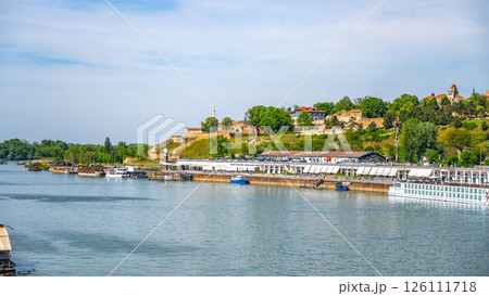 The Sava River flows peacefully through Belgrade, offering scenic views of green hills and historic architecture. Boats navigate the water while people enjoy the tranquil atmosphere. 126111718