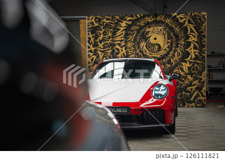 A red and white 2025 Porsche 911 GT3 922 partially covered with a wrap, parked indoors near a golden yin yang mural in a European workshop setting. 126111821
