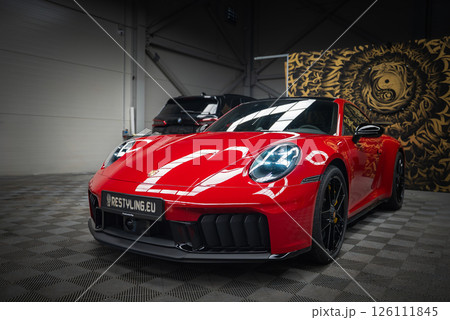 A red 2025 Porsche 911 GT3 922 with black alloy wheels sits on a checkered floor indoors. A black car and a golden yin yang wall panel are visible. A red 2025 Porsche 911 GT3 922 with black alloy wheels sits on a checkered floor indoors. A black car and a golden yin yang wall panel are visible. 126111845