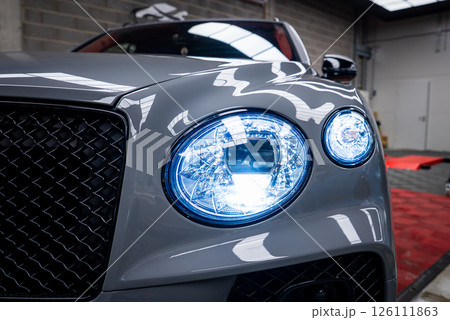 Close up view of a gray Bentley Bentayga showcasing circular LED headlights, black mesh grille, and polished surface in an indoor showroom setting. Close up view of a gray Bentley Bentayga showcasing circular LED headlights, black mesh grille, and polished surface in an indoor showroom setting. 126111863