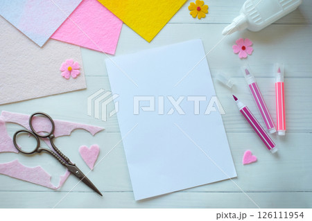 Creative flat lay composition of DIY greeting card crafting materials, including colorful felt flowers, scissors, glue, and blank paper, arranged on a white wooden background, showcasing the process 126111954