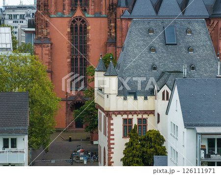 View of Frankfurt Cathedral with red sandstone facade, surrounded by traditional European buildings, greenery, and bicycles in the foreground. 126111971