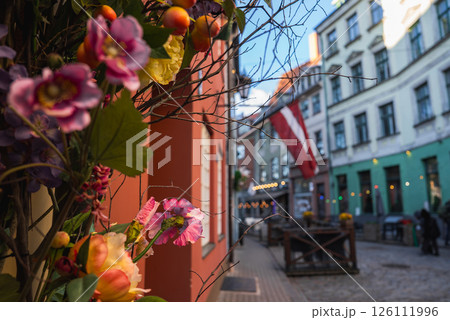 A cobblestone street in Riga's old town features pastel buildings, a Latvian flag, colorful flowers, small cafes, and string lights in a cozy setting. 126111996