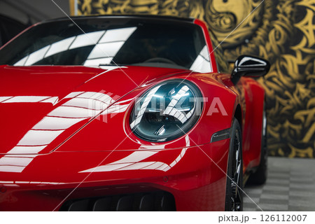 Close up of the red 2025 Porsche 911 GT3 922 front end, focusing on the left LED headlight. A gold and black mural and checkered floor are visible indoors. 126112007